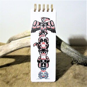 May include: A white notepad with a black and red totem pole design. The totem pole features a bird, a bear, a wolf, and a frog. The notepad has a gold spiral binding.