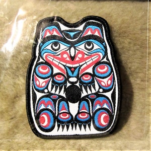 Könnte beinhalten: Ein gestickter Aufnäher in Schwarz, Weiß, Blau und Rot mit einem stilisierten indianischen Tiermotiv. Das Tier sitzt mit sichtbaren Pfoten. Das Design erinnert an einen Totempfahl.