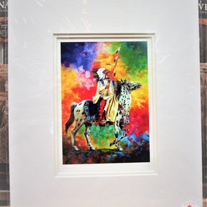 May include: A framed print of a Native American on horseback, set against a vibrant, multicolored background. The artwork features a detailed depiction of the rider in traditional attire and a spotted horse. The print is matted and framed in white.