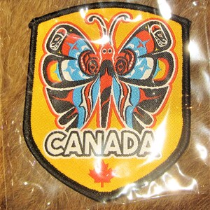 May include: A yellow felt patch in the shape of a shield with a black outline. The patch features a stylised butterfly with red, blue, black and white wings. The word "CANADA" is printed in black letters below the butterfly. A red maple leaf is in the centre of the patch.