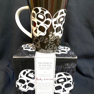May include: A black ceramic mug with a white design of two skulls. The mug is in a box with a black background and a white skull design. The box has text that reads "The Story is in the Soil" and "L'Histoire se trouve dans la Terre" by "Andy Everson".