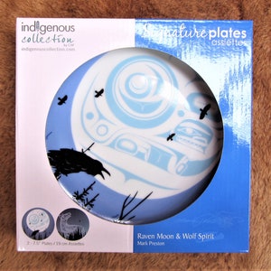 Può includere: Un piatto in ceramica bianca con un design blu e bianco di uno spirito di lupo e corvo. Il piatto ha un diametro di 19 cm e fa parte della collezione Indigenous di CAP. Il piatto è in una scatola con le parole "Signature Plates" e "Raven Moon & Wolf Spirit" stampate su di essa.