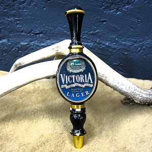 May include: A black and gold beer tap handle featuring the text "Victoria Lager" on a blue and white oval. The handle is from the Vancouver Island Brewery.