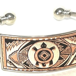 May include: A silver bangle bracelet with a rectangular copper inlay featuring a stylized turtle design in black and white. The turtle is surrounded by geometric patterns in shades of copper and white.
