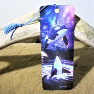 May include: A bookmark featuring two orcas leaping from the water against a starry night sky background. The bookmark is made of sturdy cardstock and has a blue tassel.