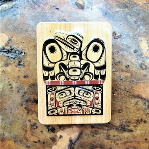 May include: A wooden rectangular box with a black and red design of a Native American eagle with a human face. The eagle is holding a smaller eagle in its talons.
