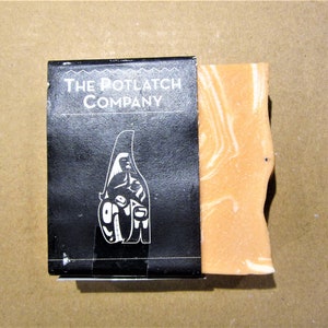 May include: A bar of soap with a peach colour and white swirls. The soap is packaged in a black box with the text "The Potlatch Company" in white.