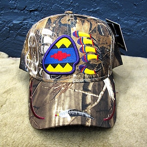 May include: Camouflage baseball cap with a colorful embroidered patch. The patch features a blue and red design with yellow and black accents. The cap also has embroidered details on the brim, including a feather and curved lines.