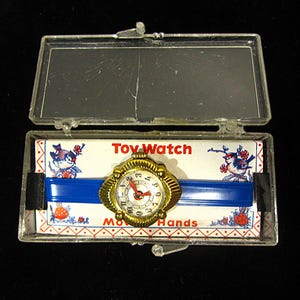 May include: A gold toy watch with a blue plastic band in a clear plastic case. The watch face has black numbers and a red hour hand. The case has a white card with the words "Toy Watch" and "Moving Hands" printed on it.