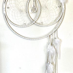 May include: White dream catcher with two connected hoops and white feathers hanging from the bottom. The dream catcher has a crystal hanging from the center.