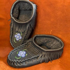 May include: A pair of black leather moccasins with fringe and beaded floral designs. The moccasins are lined with fur and have a soft, comfortable feel.