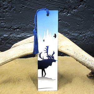 May include: A rectangular bookmark with a blue tassel. The bookmark features a blue and white landscape with a silhouette of a moose and trees. Two birds are flying in the sky. The bookmark is made of cardstock.
