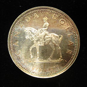 May include: A silver Canadian dollar coin featuring a depiction of a mounted Royal Canadian Mounted Police officer. The coin has the text "CANADA" and "ONE DOLLAR" around the edge, and the years "1970" and "1979" at the bottom.
