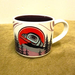 May include: White ceramic mug with a colorful design of a stylized bird in a red circle, surrounded by trees and a mountain range. The mug has a purple interior.