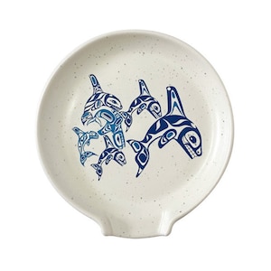 May include: A white ceramic spoon rest with a blue and white design of orcas. The orcas are stylized in a traditional Northwest Coast Indigenous art style.