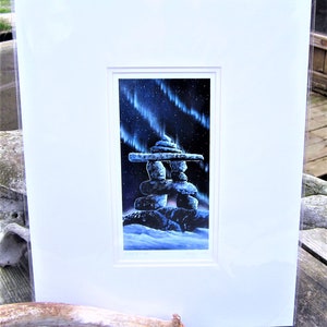 Op de afbeelding: Een ingelijste print van een stenen inukshuk-beeldhouwwerk tegen een achtergrond van het noorderlicht, een levendig spektakel van groene en blauwe lichten in de nachtelijke hemel.