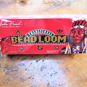 Op de afbeelding: Een rode doos met de woorden "John Bead Established 1954" en "Traditional Bead Loom" erop gedrukt. De doos heeft een illustratie van een indiaan met een hoofdtooi en een kralenpatroon.