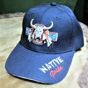 May include: A navy blue baseball cap with an embroidered image of a Native American headdress and a buffalo skull. The cap has white embroidery that says "NATIVE Pride" on the front.