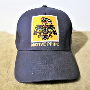 May include: A navy blue baseball cap with a yellow square patch featuring a colorful Native American eagle design. The text "NATIVE PRIDE" is embroidered below the patch in rainbow colors.