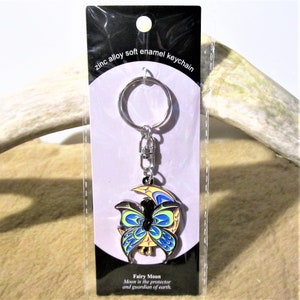 Può includere: Un portachiavi d'argento con un ciondolo smaltato blu e giallo di una fata con le ali seduta su una luna crescente. Il ciondolo ha il testo "Fairy Moon Moon is the protector and guardian of earth."