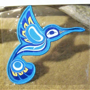 Può includere: Una toppa ricamata di colibrì blu e gialla con un occhio bianco. Il colibrì è in posizione di volo con le ali spiegate.