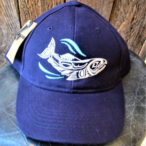 May include: A navy blue baseball cap featuring a white and blue embroidered salmon design. The salmon is styled with a traditional Northwest Coast design.