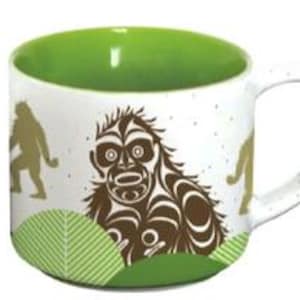 May include: White ceramic coffee mug with a green interior and a brown and white graphic of a stylized Sasquatch with a green and white background.