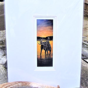 Op de afbeelding: Een ingelijste print van een schilderij dat twee elanden laat zien die in een meer staan ​​bij zonsondergang. De elanden zijn gesilhouetteerd tegen de oranje en gele lucht. De print is getiteld "Evening Shadows - Nature's Grace".