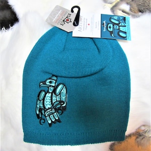 May include: Teal blue knit toque with a black and blue embroidered raven design. The toque is made in Canada and features the "Oscar & Co." and "Artis" labels.