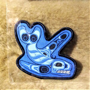 May include: A blue and black embroidered patch depicting a stylized owl with a fish in its beak. The owl is surrounded by intricate designs and patterns.