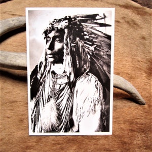 May include: Black and white photograph of a Native American man wearing a traditional headdress with feathers. He is wearing a beaded necklace and a fringed garment.