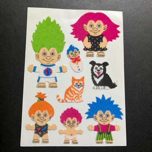 Può includere: Un foglio di adesivi colorati con vari personaggi troll, tra cui un troll verde con una camicia blu, un troll rosa con un vestito a pois neri e bianchi, un troll blu con pantaloni verdi, un troll dai capelli rossi con un costume da bagno rosa, un troll rosa con un pannolino bianco, un gatto, un cane e un bambino troll.