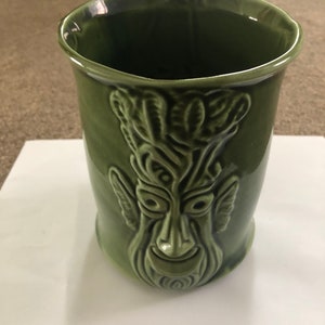 Może przedstawiać: Zielony kubek ceramiczny z rzeźbioną na przodzie szczegółową twarzą. Twarz ma szerokie usta, duże oczy i gęstą brodę.