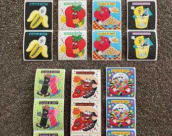 May include: A collection of vintage "Scratch & Sniff" stickers featuring various food and character designs. The stickers include images of bananas, strawberries, apples, and other playful illustrations. Each sticker has a white border and the words "Scratch & Sniff".