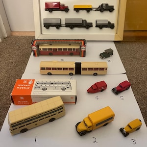 以下が含まれることがあります： 赤、黄色、グレーなどの様々な色のバス、トラック、路面電車など、ヴィンテージの玩具車両のコレクション。一部は額縁に、その他は白い表面に配置されています。箱入りのモデルも含まれています。