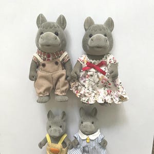 Pu&ograve; includere: Quattro statuette di ippopotami grigi. Due ippopotami adulti sono vestiti con una camicia a quadri con salopette beige e un vestito floreale con un fiocco rosso. Due ippopotami pi&ugrave; piccoli indossano salopette gialle con una tasca arancione e un vestito a righe blu.