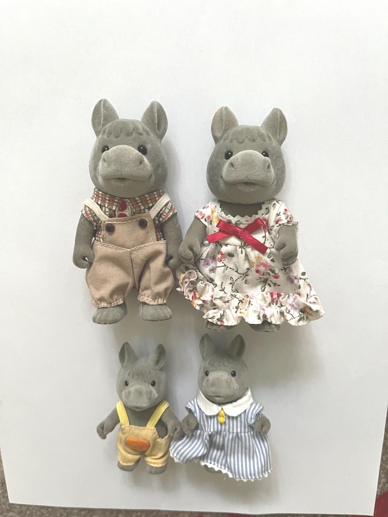 Pu&ograve; includere: Quattro statuette di ippopotami grigi. Due ippopotami adulti sono vestiti con una camicia a quadri con salopette beige e un vestito floreale con un fiocco rosso. Due ippopotami pi&ugrave; piccoli indossano salopette gialle con una tasca arancione e un vestito a righe blu.