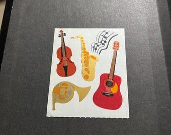 May include: A set of five musical instrument stickers featuring a violin, saxophone, french horn, guitar, and musical notes. The stickers are on a white background.