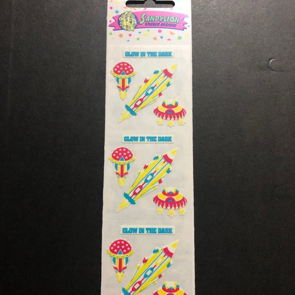 Può includere: Un foglio di adesivi che brillano al buio con un razzo, un UFO e un oggetto rosso, bianco e blu con stelle. Gli adesivi sono di Sandylion Sticker Designs.
