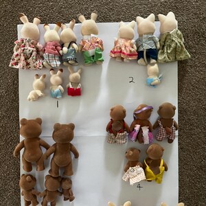May include: A collection of flocked animal figurines, including rabbits and bears, arranged on a white sheet. The figures are shown from the back, wearing various fabric outfits like dresses, shirts, and overalls. Some figures are labelled with handwritten numbers.