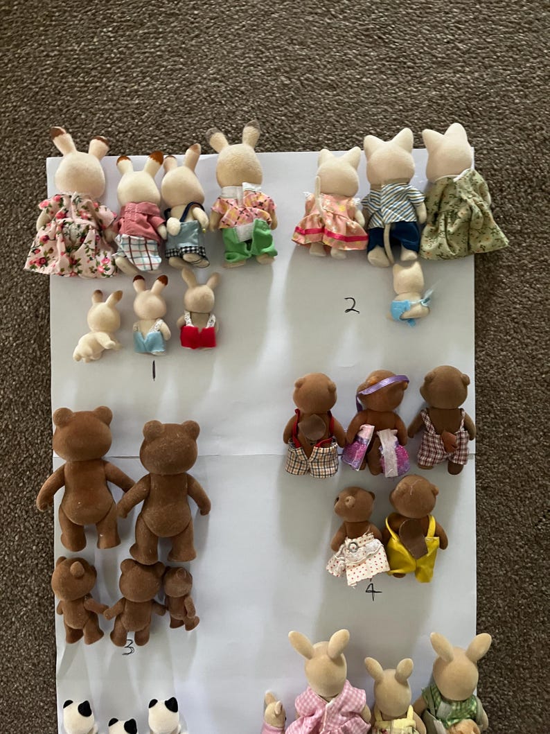 May include: A collection of flocked animal figurines, including rabbits and bears, arranged on a white sheet. The figures are shown from the back, wearing various fabric outfits like dresses, shirts, and overalls. Some figures are labelled with handwritten numbers.