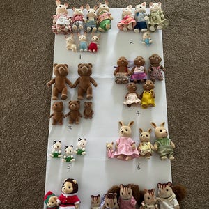 May include: A collection of flocked animal figurines arranged in eight numbered groups on a white surface. The set includes various rabbits, bears, and squirrels wearing fabric clothing, with some smaller baby figures and some larger adult figures.