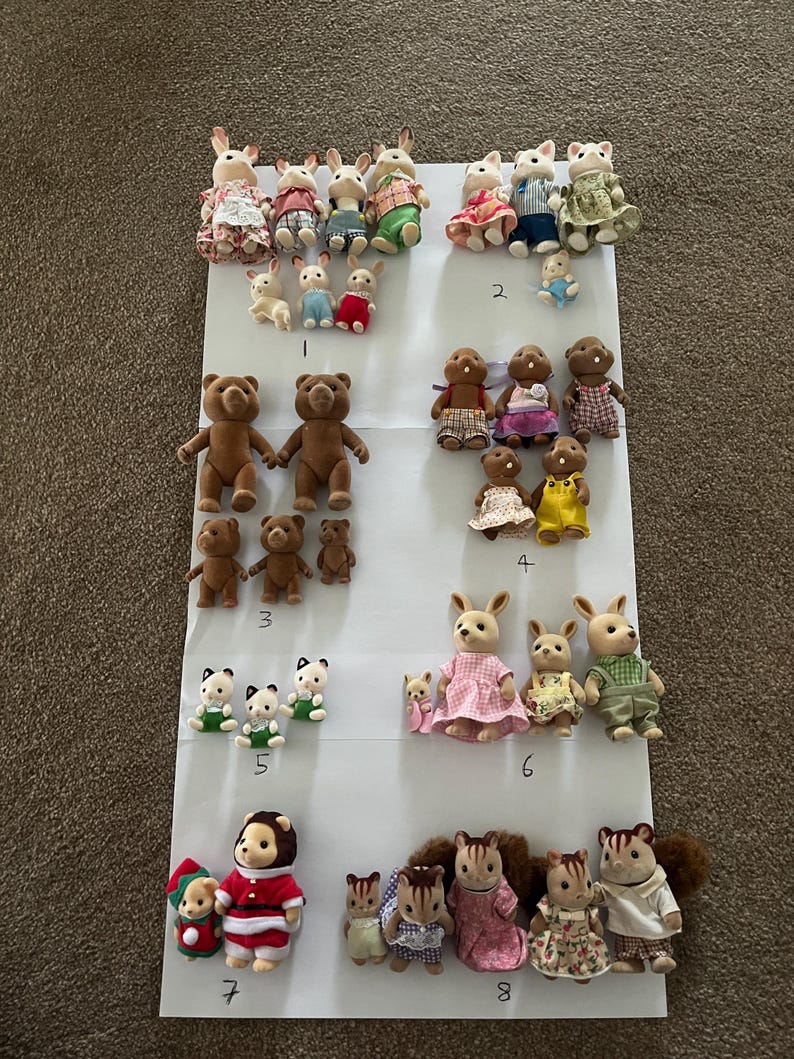 May include: A collection of flocked animal figurines arranged in eight numbered groups on a white surface. The set includes various rabbits, bears, and squirrels wearing fabric clothing, with some smaller baby figures and some larger adult figures.