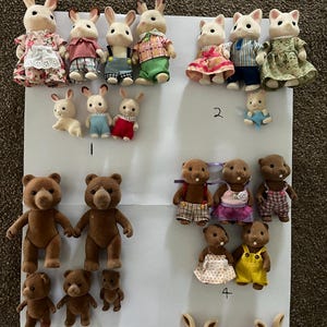 May include: A collection of flocked animal figurines including rabbits, bears, and cats dressed in various fabric outfits. The figures are arranged in groups on a white sheet, with some handwritten numbers visible near specific sets of characters.