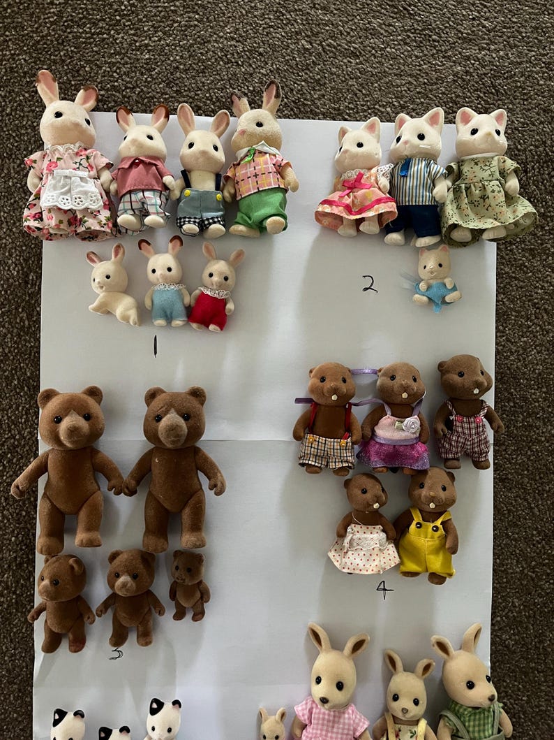 May include: A collection of flocked animal figurines including rabbits, bears, and cats dressed in various fabric outfits. The figures are arranged in groups on a white sheet, with some handwritten numbers visible near specific sets of characters.