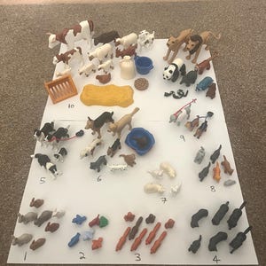 May include: A collection of plastic toy farm and zoo animals, including cows, sheep, lions, pandas, and dogs, arranged on a white surface. Various accessories like a blue bucket and hay bale are also present. The toys are numbered 1-10.