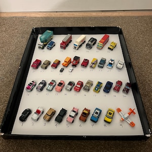 Può includere: Una collezione di veicoli giocattolo in miniatura, tra cui camion, auto e un aereo, disposti su una superficie bianca all'interno di un vassoio nero. I giocattoli sono numerati e variano di colore, come rosso, blu, giallo e verde.
