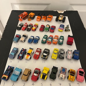 Può includere: Una collezione di 26 auto e camion in miniatura, tra cui un veicolo da costruzione, un monster truck e una varietà di auto sportive. Le auto sono disposte in file su una superficie bianca e numerate da 1 a 26.