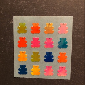 May include: A sheet of 16 holographic bear stickers in various colours, including orange, pink, blue, green, and yellow. The bears have a simple design with black eyes and a small smile.