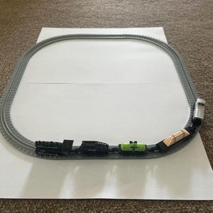 May include: A toy train set featuring a black locomotive, coal car, green tanker car, and a car carrying wooden logs. The train runs on a grey track, forming an oval. The set is on a white surface.